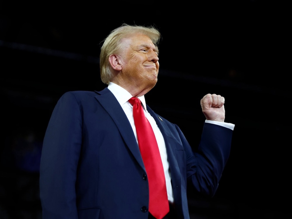 A 78-year-old light-skinned man with styled blondish hair stands at a podium and holds one fist in the air.
