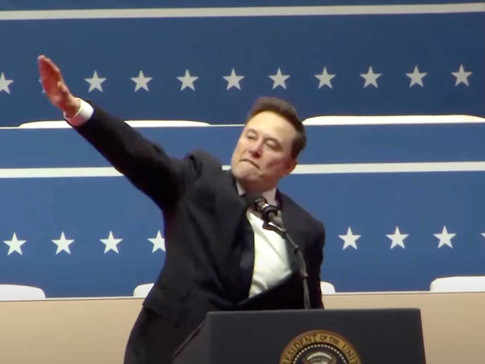 A 53-year-old light-skinned man wearing a dark suit stands at a podium, his right arm extended in a Nazi salute.