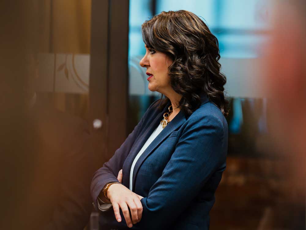 A 53-year-old light-skinned woman with shoulder length dark brown hair and wearing a blue suit stands with arms crossed.