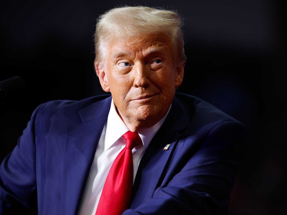 Donald Trump, a light-skinned man with blow-dried light hair, smiles while wearing a blue suit and red tie.
