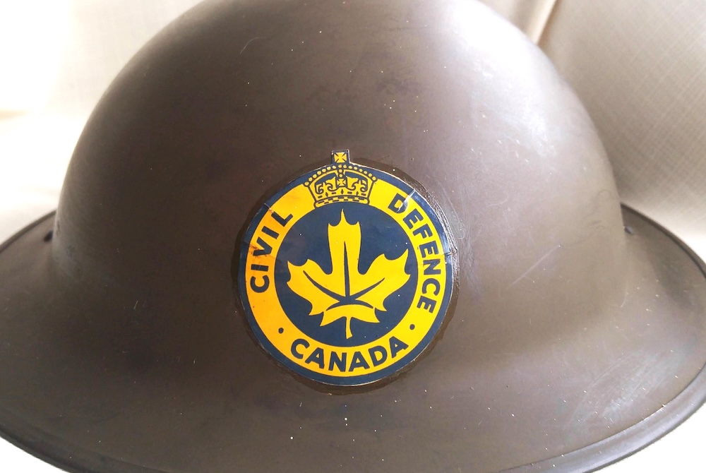 A dark brown helmet with a round yellow logo featuring a maple leaf and the words 'Civil Defence Canada.'