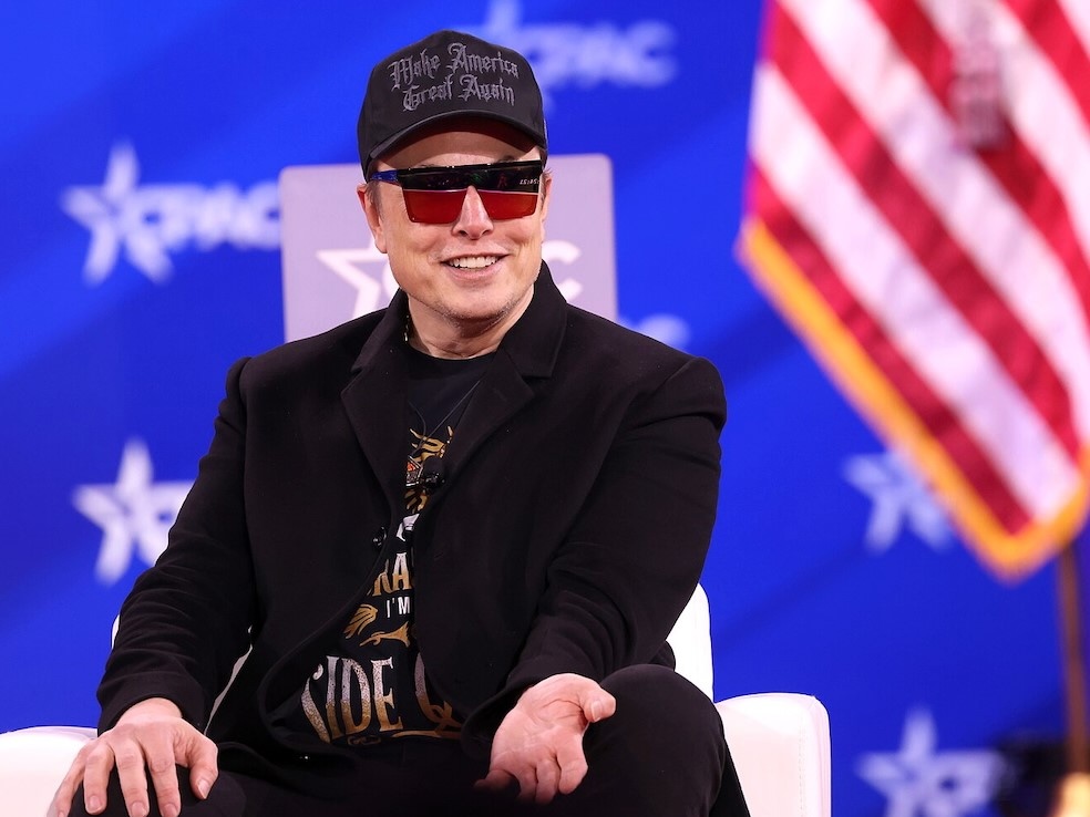 Elon Musk, a 53-year-old light-skinned man, sits on a stage. He wears sunglasses, a black overcoat and a black ball cap that says &lsquo;Make America Great Again&rsquo; in gothic script.
