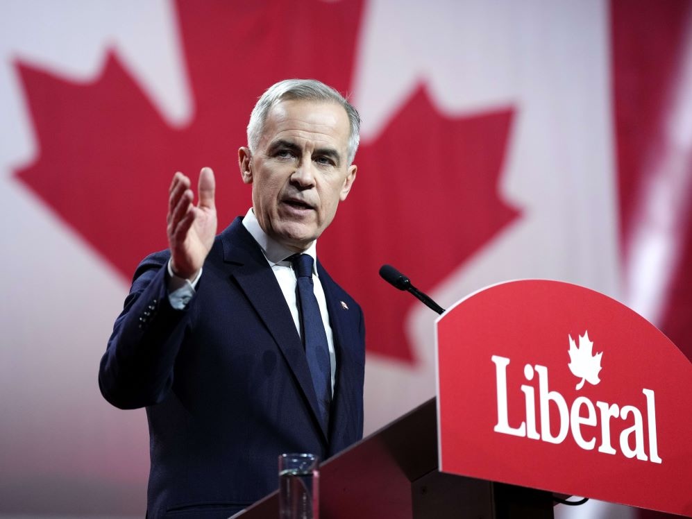A 59-year-old man with light skin and short grey hair, wearing a navy blue suit and tie, speaks at a podium behind a 'Liberal' sign, with a large Canadian flag behind him.