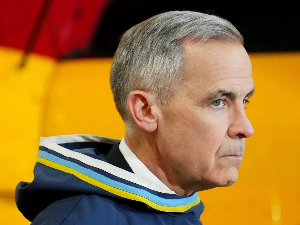 Mark Carney is shot in profile. He has medium skin and short grey hair. He is facing the right of the frame and wearing a hooded garment with a white, navy, light blue and yellow band. He is standing against a yellow background.