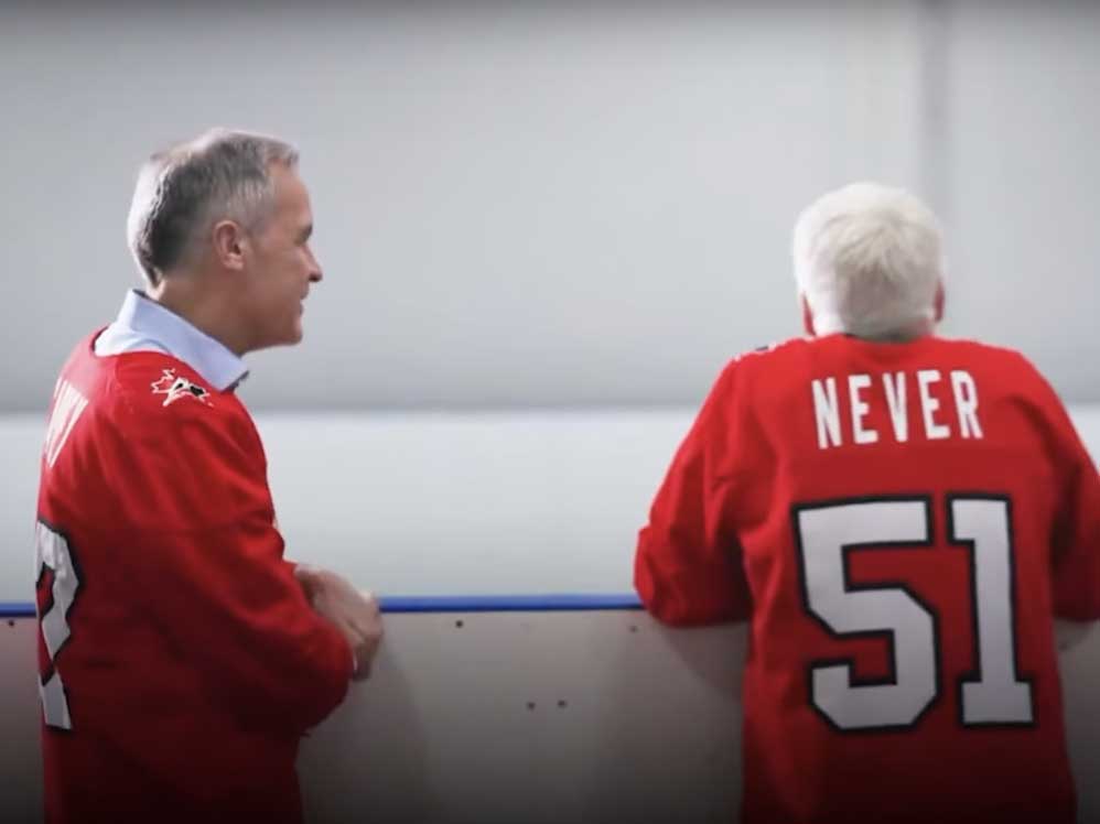 Two men lean on hockey boards, both older, grey-haired, light-skinned and wearing red hockey jerseys.