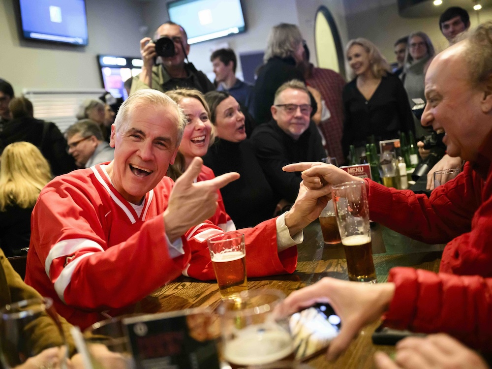 Mark Carney, wearing a red hockey jersey, smiles and gestures in a crowded pub, a beer on the table in front of him.