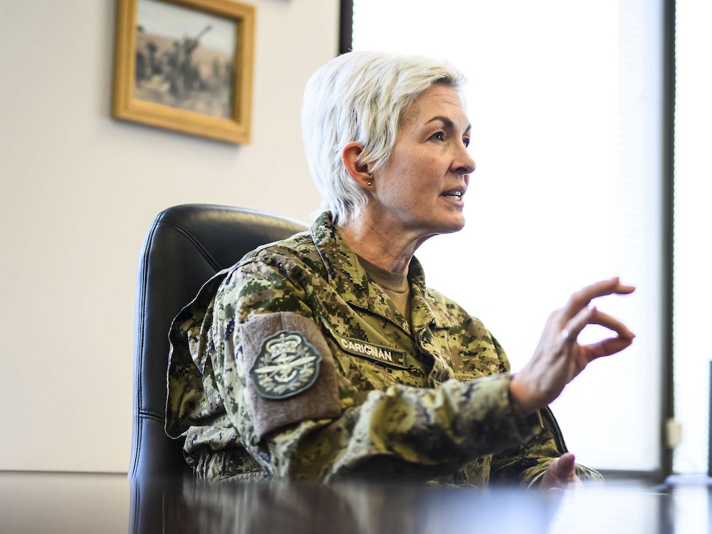 Jennie Carignan has short, light grey hair and is wearing green military fatigues. She is seated in a black office chair across a dark table in an executive office. Her hand is raised mid-speech.