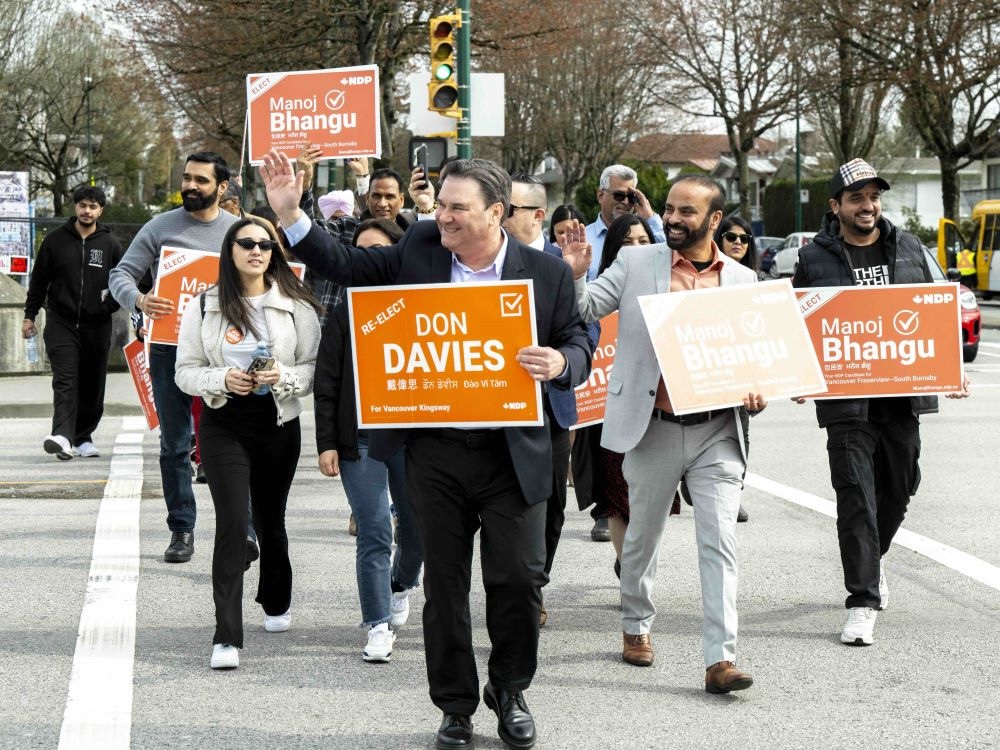 Don Davies has short brown hair and pale skin. He is wearing a dark suit and crossing a residential street with a crowd of people holding orange signs for New Democratic Party candidates. Davies is smiling and waving, looking to the left of the frame.