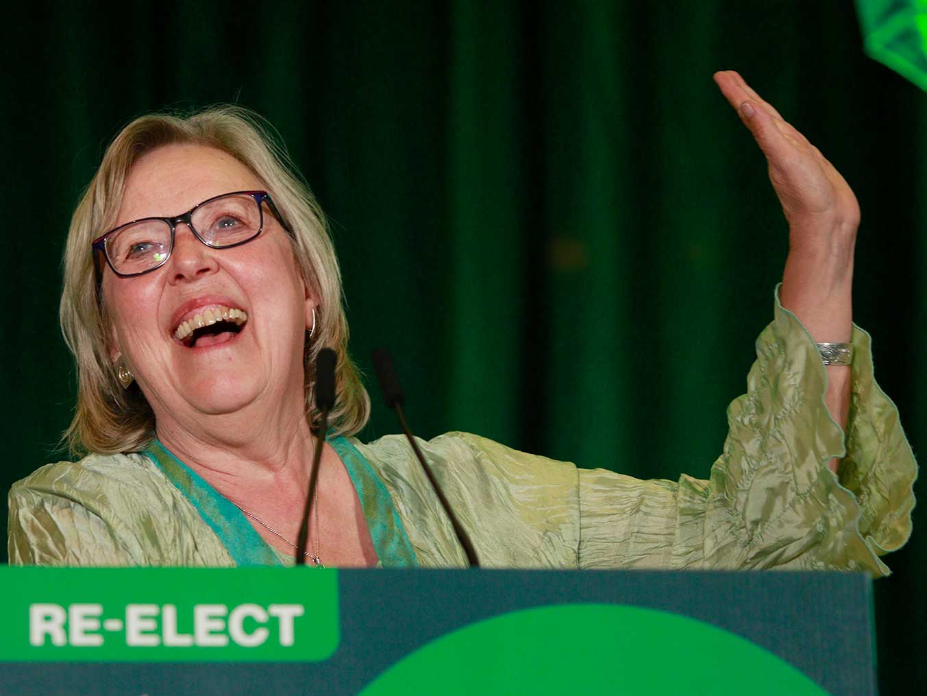 A 70-year-old woman with light-coloured hair, wearing a light green shirt, smiles and holds up one hand.