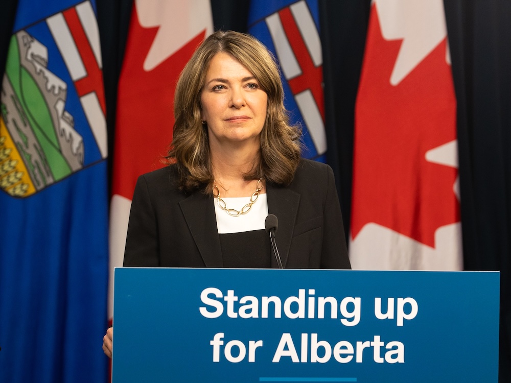 Danielle Smith, a 54-year-old woman, stands at a lectern with a sign that says &lsquo;Standing up for Alberta.&rsquo; She&rsquo;s wearing a black jacket, white shirt and gold necklace.