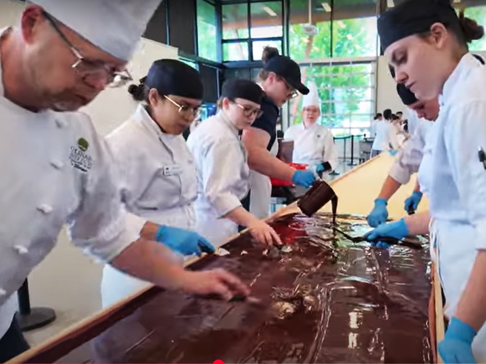 People in white chef’s garb pour chocolate as part of the preparation of a giant Nanaimo bar.