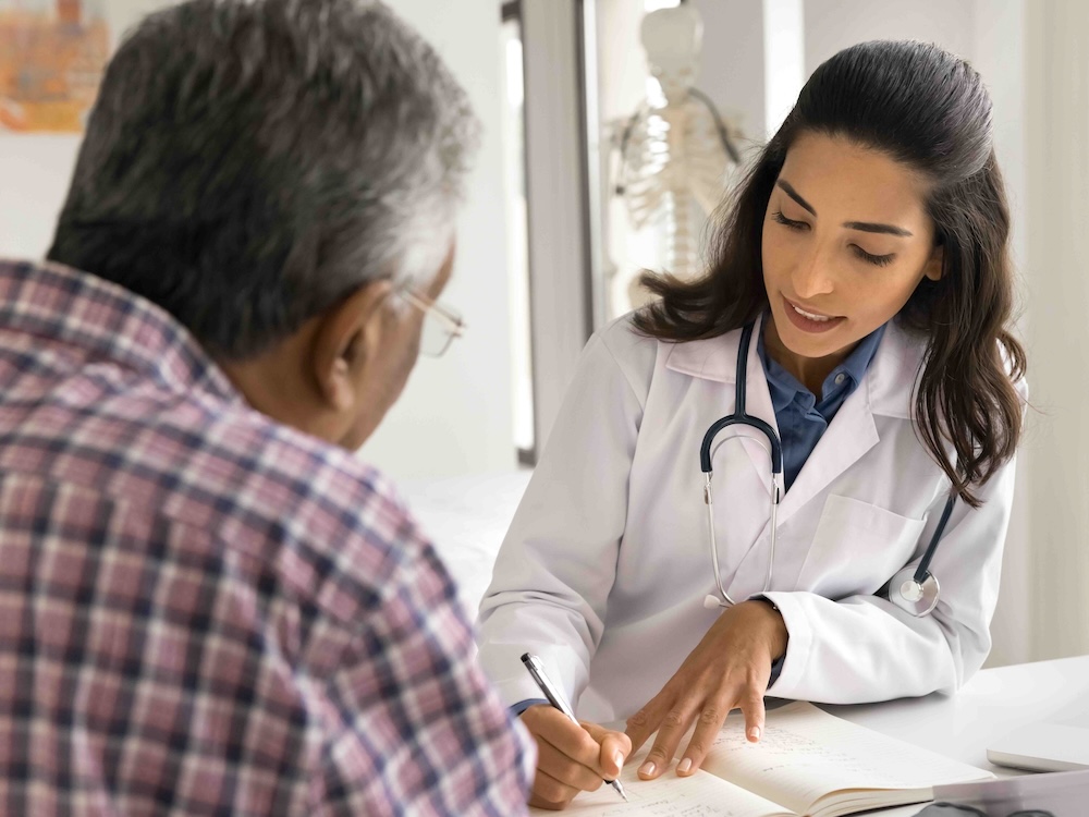 A woman in a white lab coat with a stethoscope around her neck is seated at a desk, writing in a notebook. She has dark hair and a medium skin tone. In front of her is a man with his back turned to the camera. He has dark hair and a plaid shirt.