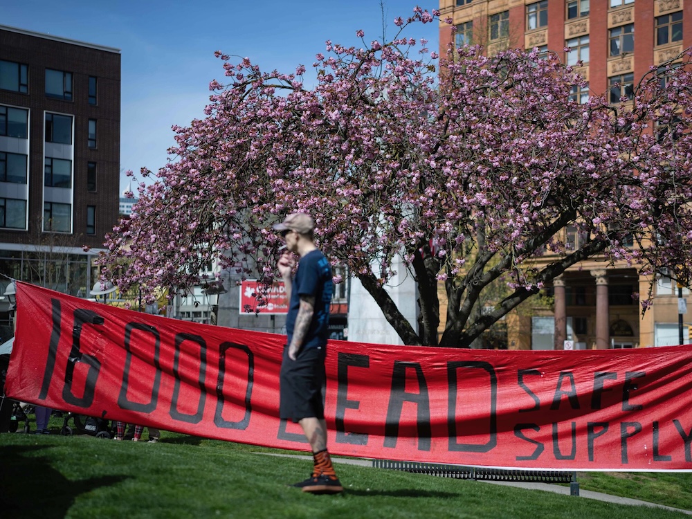 A person in shorts and a T-shirt stands on grass in front of a red banner that says ‘16,000 Dead. Safe Supply.’