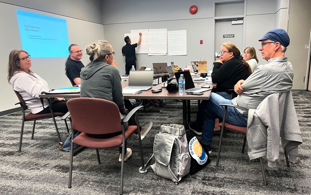 Six people sit around a conference table littered with papers, laptops and water bottles while one man writes on a whiteboard in the background.