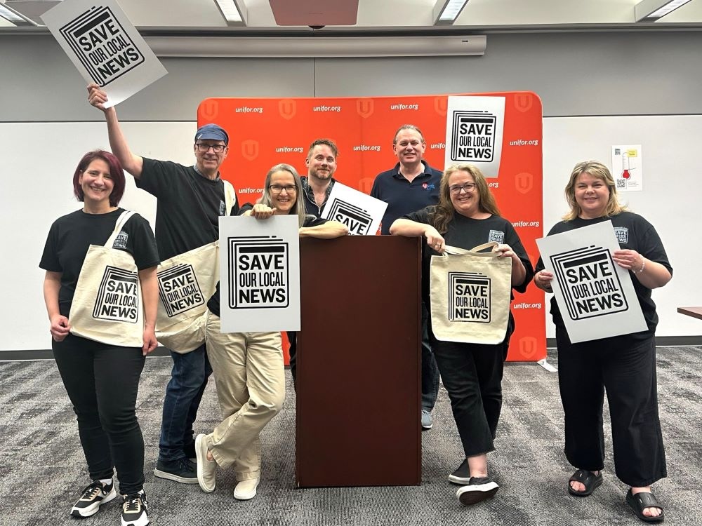 Seven people stand around a lectern holding signs and tote bags that read ‘Save Our Local News.’