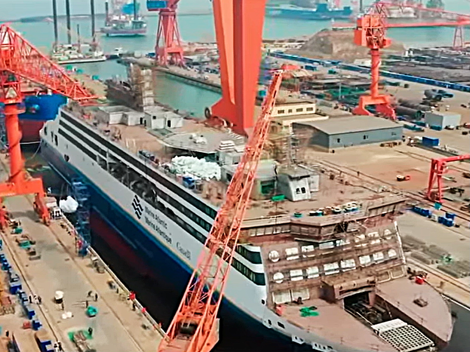 An aerial view of a large ship with a blue hull and white superstructure under construction in a shipyard. 