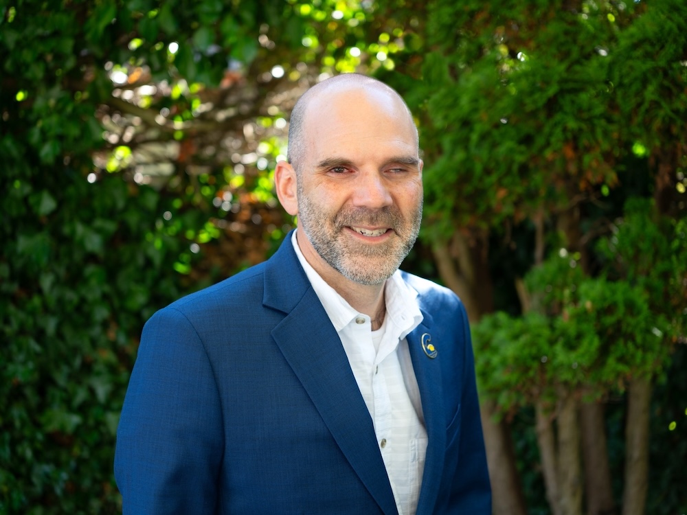 A 40-ish light-skinned man, balding and with a close-cropped beard, stands outdoors.