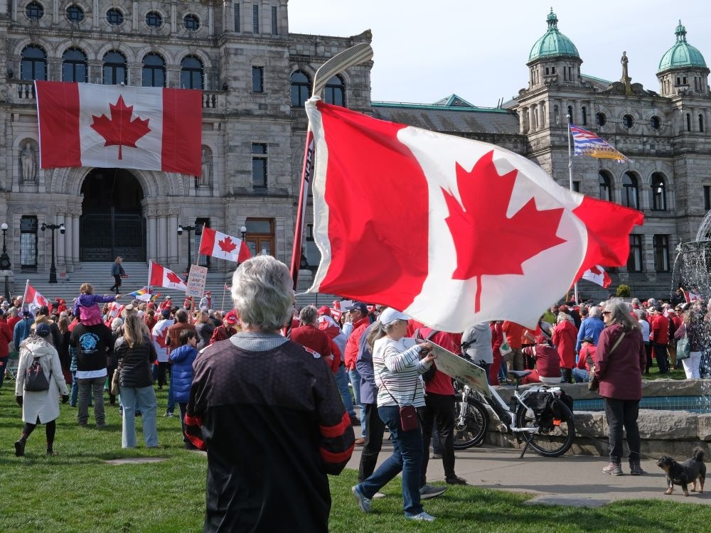 A man with salt-and-pepper hair is seen from behind hoisting a Maple Leaf flag on a hockey stick. In the distance are other crowd members, Canadian flags and the B.C. legislature building.