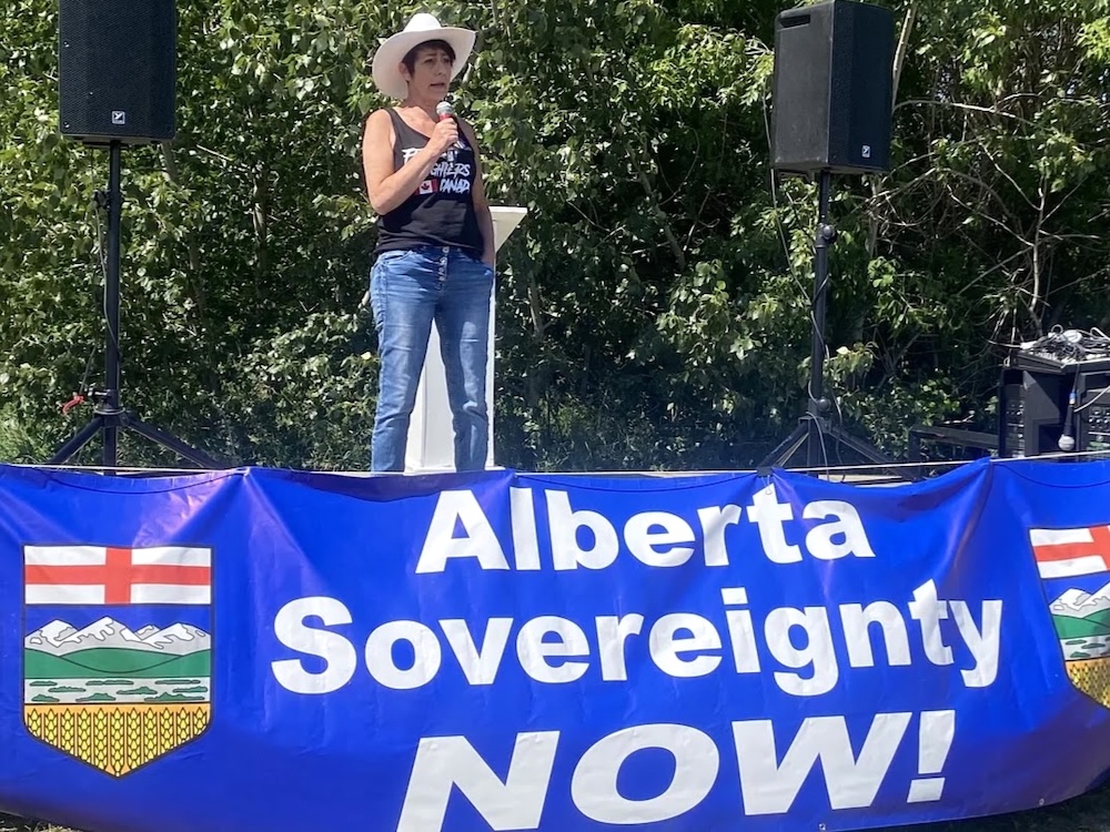 A light-skinned woman in a white cowboy hat, jeans and a black tank top stands on a stage. In front of her is a large banner that says ‘Alberta Sovereignty NOW!’