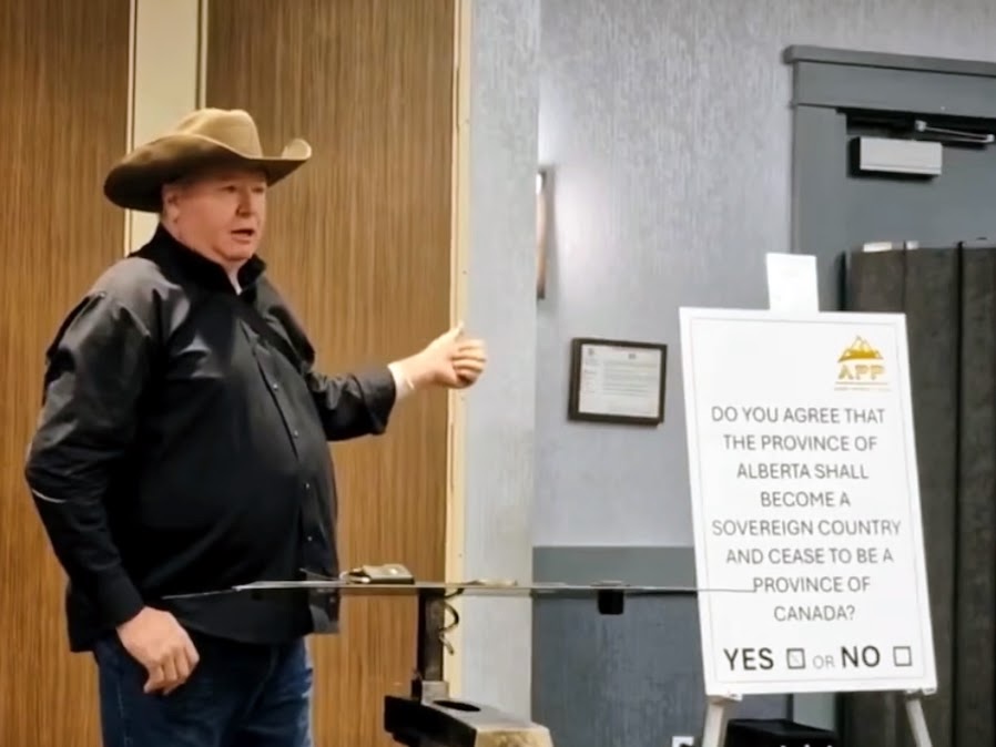 A large man with light skin tone, wearing a black shirt and brown cowboy hat, stands beside a sign with writing on it.
