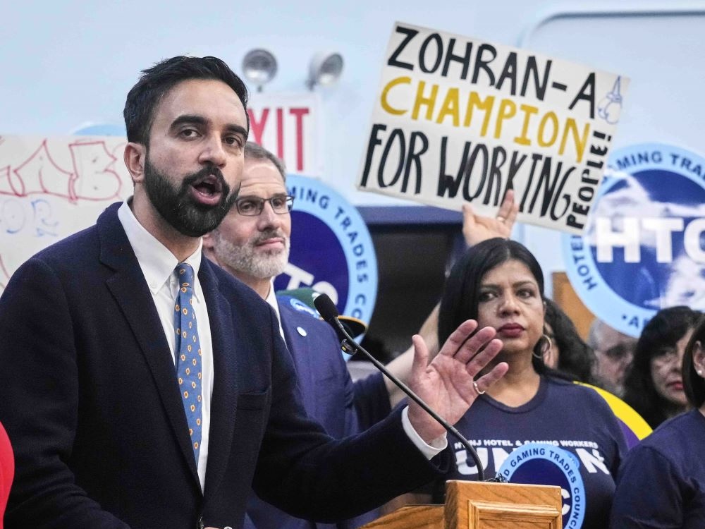 Zohran Mamdani has short dark hair and a beard. He is wearing a dark suit over a white shirt and speaking into a microphone at a wooden lectern during a rally. Behind him, people are holding signs including a handwritten one that reads &lsquo;Zohran &mdash; a champion for working people.&rsquo;