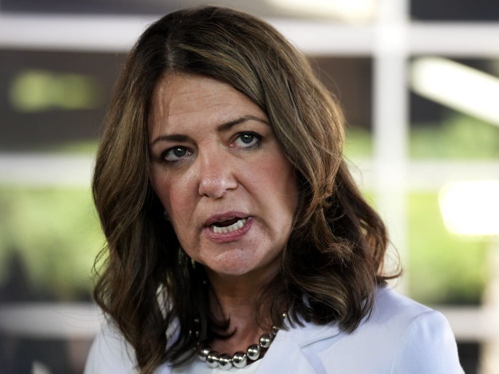 A middle aged woman with dark brown curly hair, Alberta Premier Danielle Smith, is wearing a white blouse and a necklace. She looks to her side with her eye brows furrowed.