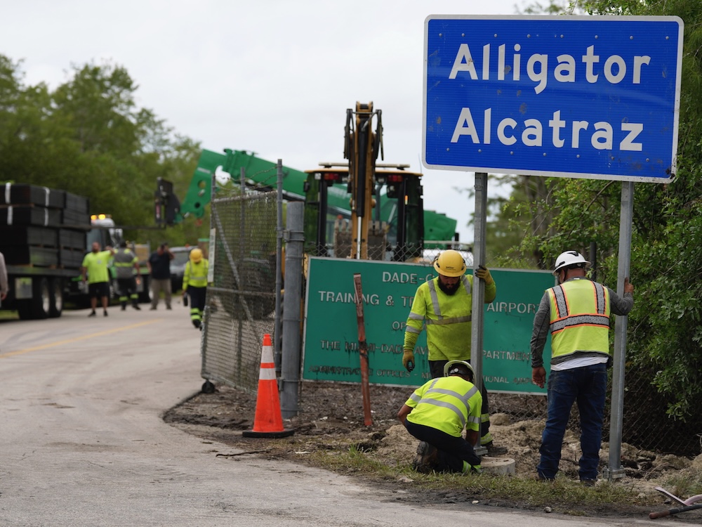 Workers wearing bright yellow jackets and pants and hard hats install a road sign that reads &lsquo;Alligator Alcatraz&rsquo; at the side of a road.