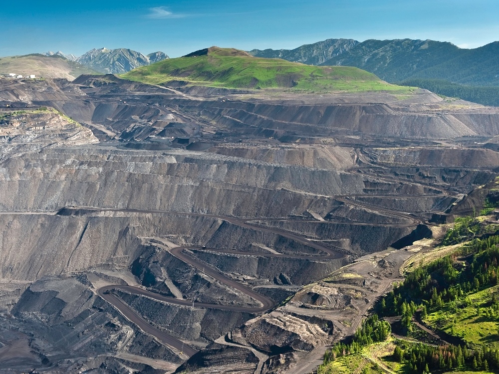 A large, tiered open-pit coal mine, with blue sky above and mountains in the background.