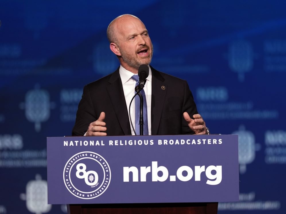 A light-skinned bald man wearing a dark suit speaks from a podium.