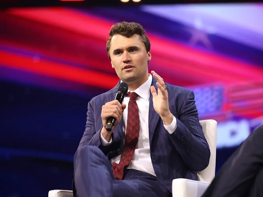 A man with light skin and short wavy brown hair wearing a suit and tie is seated in a chair, holding and speaking into a microphone.