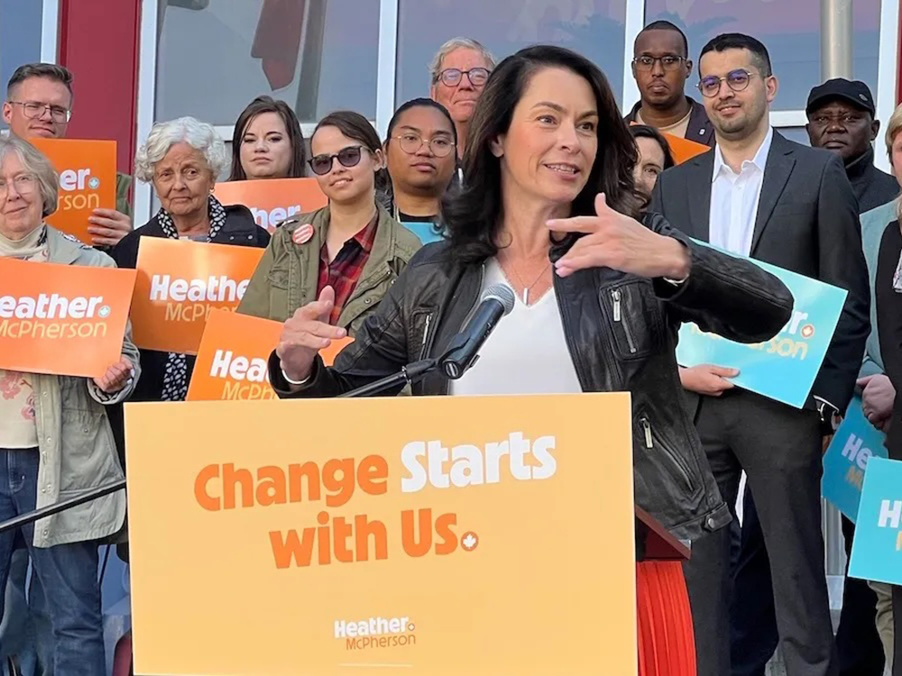 Heather McPherson, a 53-year-old light-skinned woman wearing a black leather jacket, is backed by supporters as she speaks at a podium.