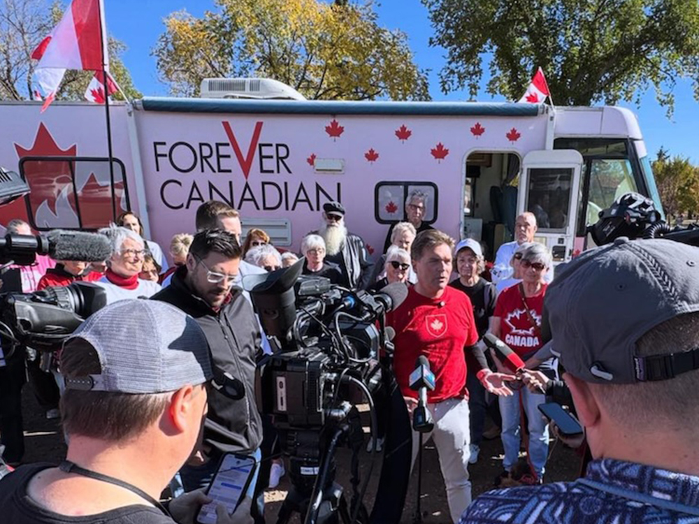 A man in a red shirt speaks outdoors to a large group of reporters. Behind him are supporters and an RV with red maple leafs and the words &ldquo;Forever Canada.&rdquo;