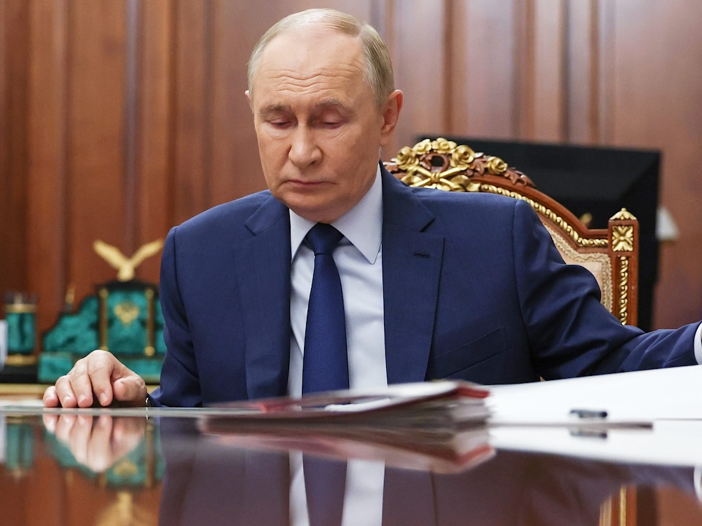 Vladimir Putin is seated on an ornate chair at a polished desk in an office with brown walls and flags in the background. He is looking down at the documents before him. He has thin light brown hair and a pale skin tone and is wearing a navy suit.