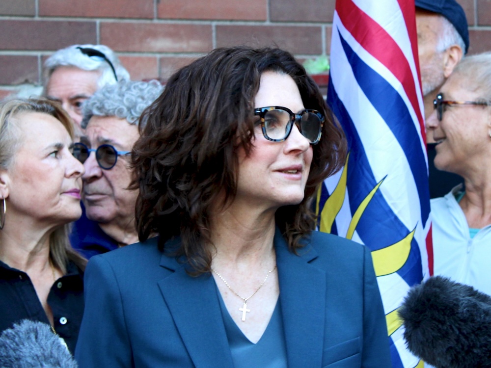 A light-skinned woman with shoulder-length brown hair stands with microphones in front of her. A group of people and the British Columbia flag can be seen behind her.