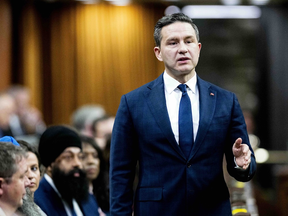 Pierre Poilievre stands in the House of Commons as other MPs look on.