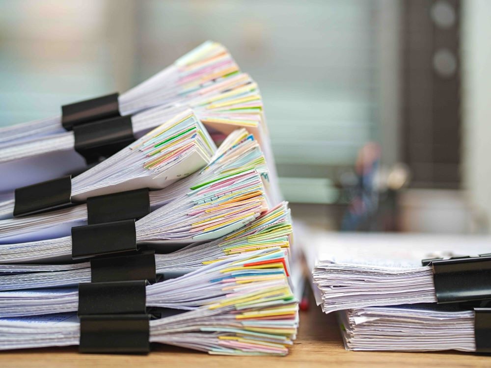 Two stacks of paper documents sit beside each other on a wooden desk. The stack of white papers with black clips on the left is annotated with colourful markers. It is taller than the stack on the right.
