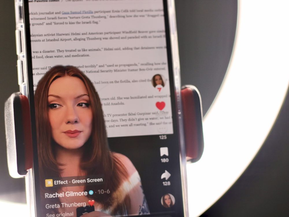 A phone shows a still from a TikTok video that shows a woman with long auburn hair speaking with an image showing the text of a news story. A ring light can be seen behind the phone.