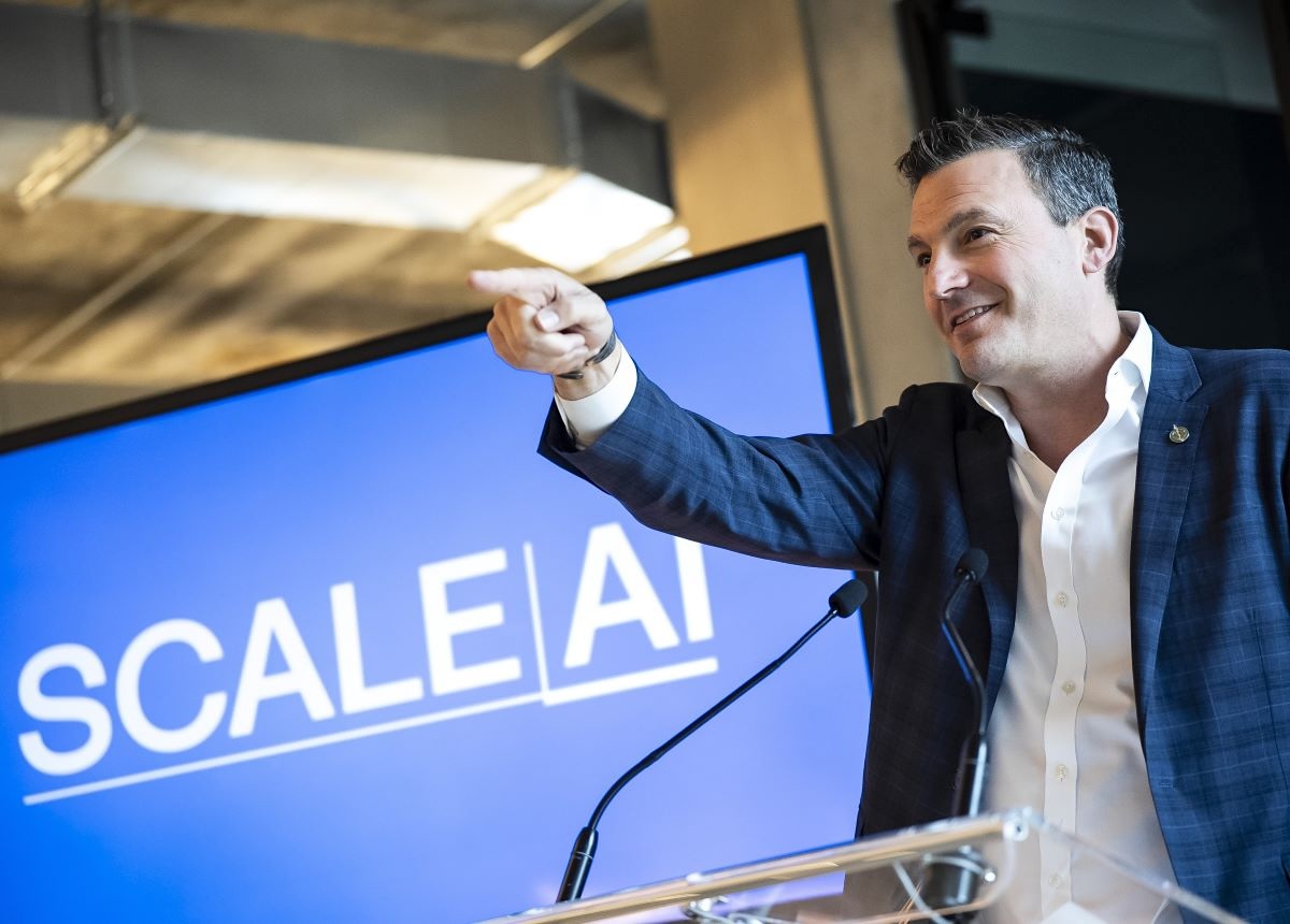 A dark-haired middle-aged man in an open-collar white shirt and blue plaid sports coat points and smiles in front of a video screen that says &lsquo;Scale AI.&rsquo;