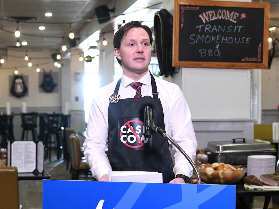 A 37-year-old light-skinned man stands at a podium in a restaurant, wearing a blue apron that saying &ldquo;Cash cow.&rdquo;