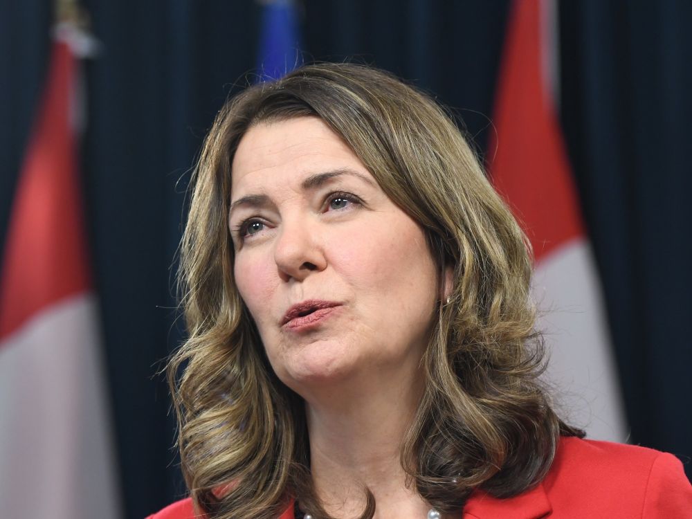 A close-up photo of Danielle Smith, a middle-aged white woman with shoulder-length brown hair, with three multicoloured flags out of focus behind her.