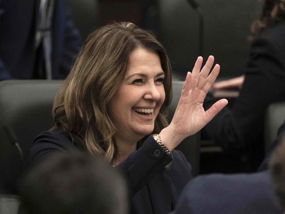 Danielle Smith has wavy shoulder-length brown hair and a light skin tone. She is seated on a black leather chair in an indoor setting, smiling and waving.