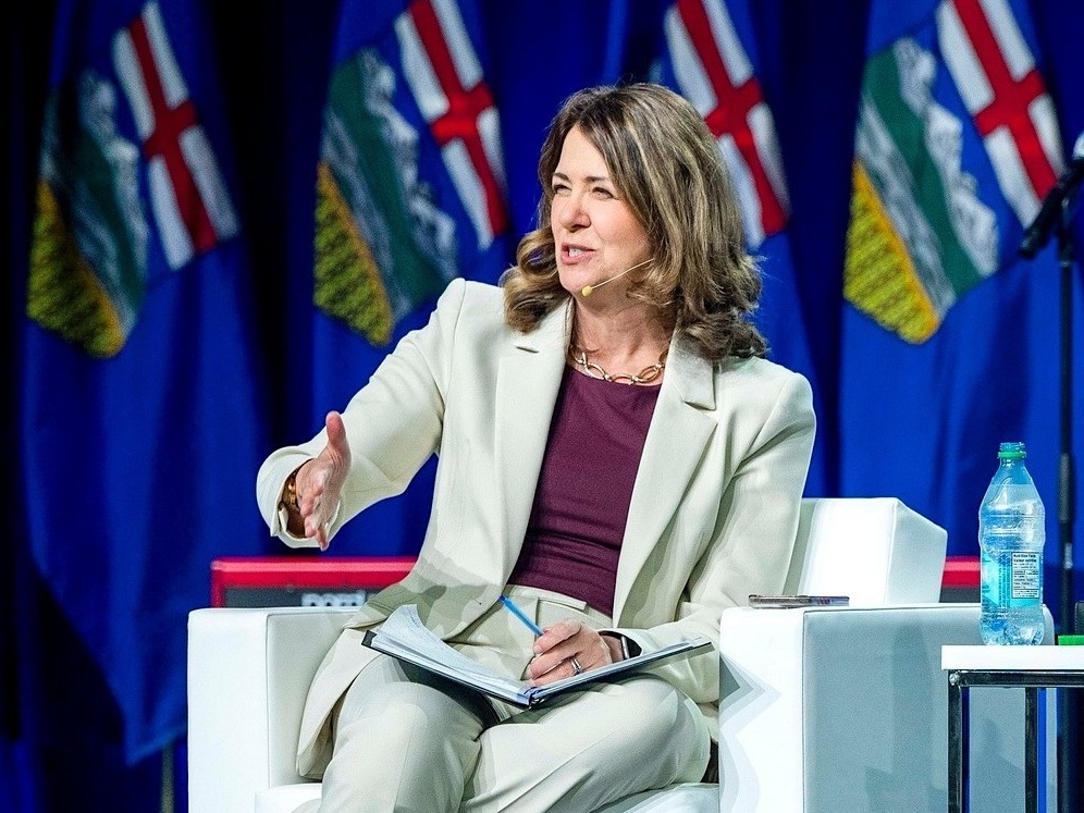 Danielle Smith, wearing a cream-coloured pantsuit and seated in a white chair, gestures with one hand while speaking.