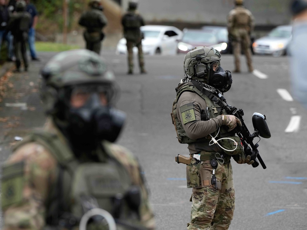 Two Immigration and Customs Enforcement agents stand on a street. They are wearing body armour, helmets and khaki-coloured uniforms. One carries a long weapon that looks like a gun but is likely a pepper ball launcher.
