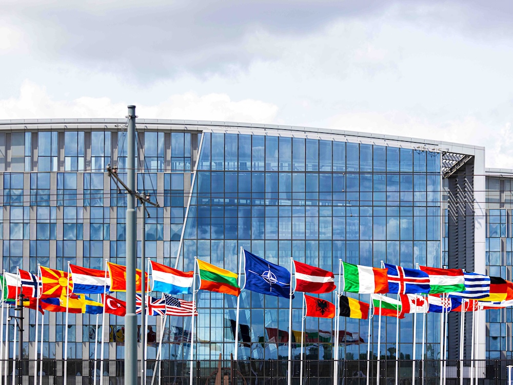 International flags stand on white poles with the blue NATO flag in the middle. They are in front of a tall glass building on an overcast day.