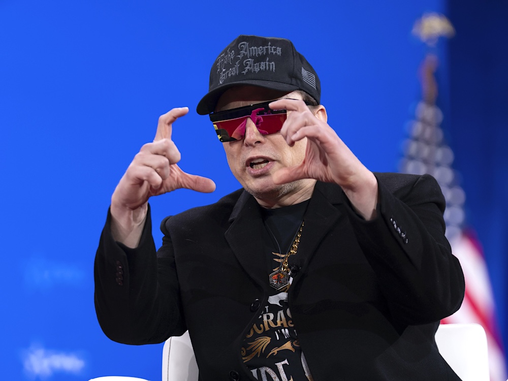A man in his 50s wears a black cap, mirrored sunglasses, a black T-shirt and black blazer as he gestures while sitting in a chair on a stage.