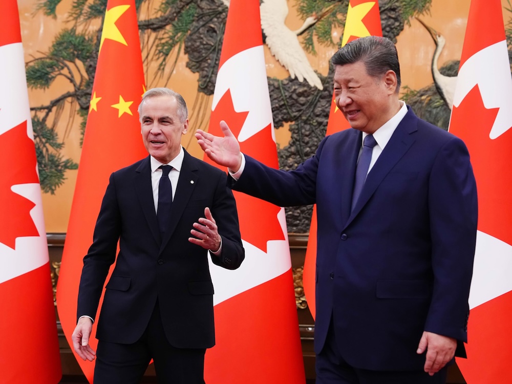 Mark Carney, in a black suit, stands beside Xi Jinping in a blue suit, in front of Chinese and Canadian flags.