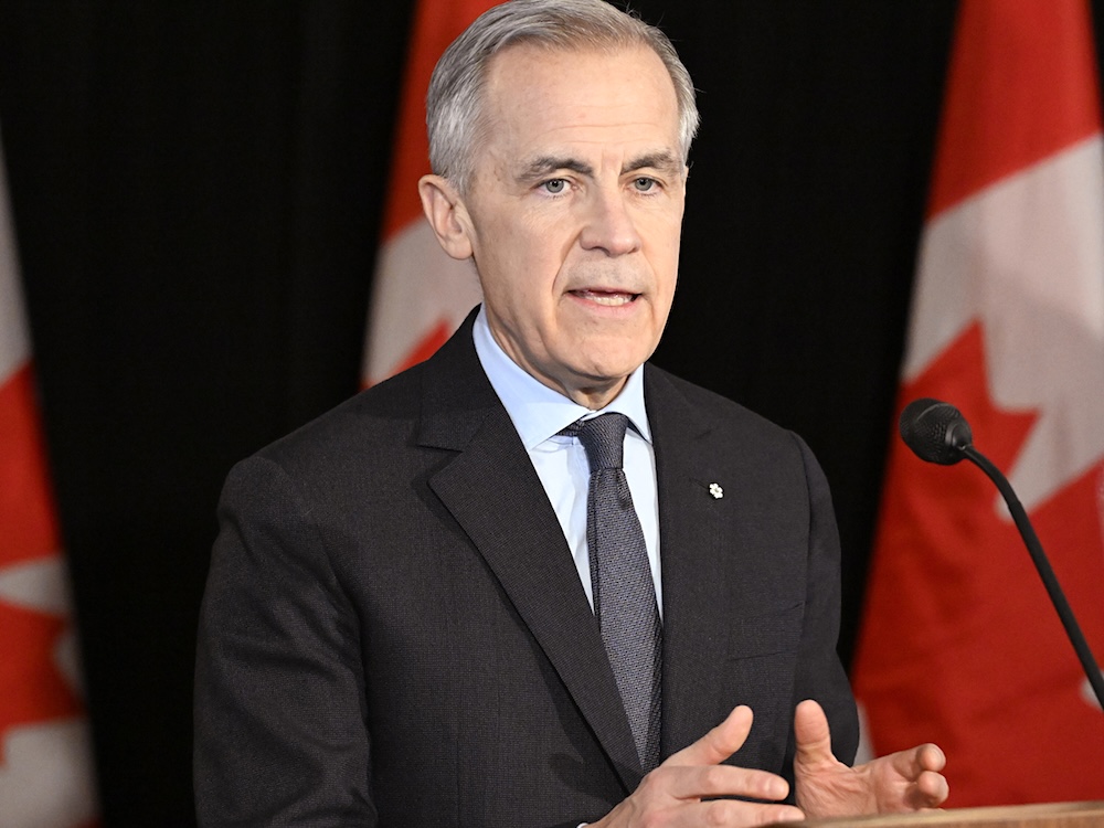 Mark Carney gives a speech. He is in front of Canadian flags, behind a lectern.