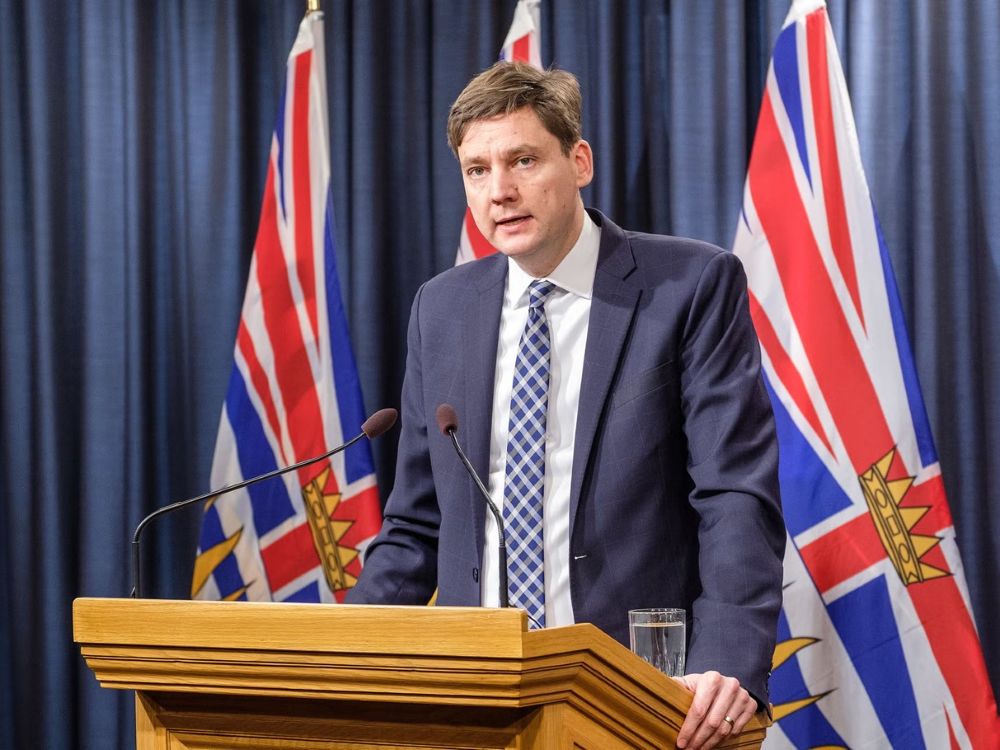 A light skinned middle-aged man in a dark blue suit and striped tie, B.C. Premier David Eby, stands at a microphoned podium with the flags of British Columbia draped behind him.