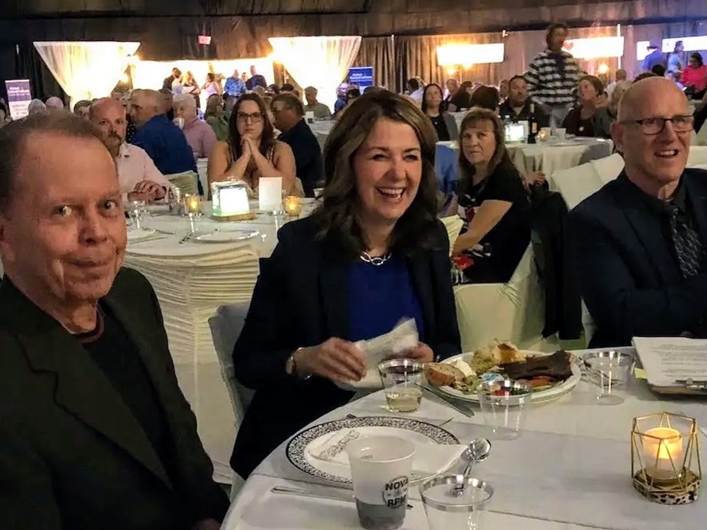 Danielle Smith, a woman with light skin and shoulder-length brown hair wearing a black blazer, sits laughing at a table at a dinner. On either side are two light-skinned, older men dressed in black.