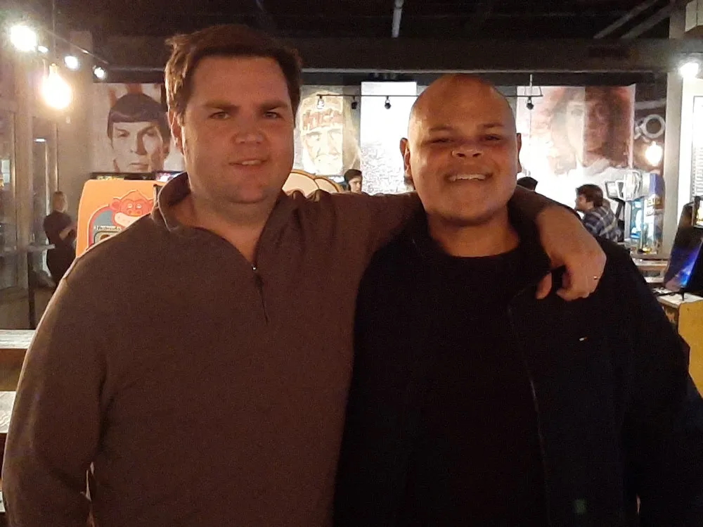 Two 40ish men stand arm and arm, one light-skinned with short brown hair and the other with medium-toned skin and a shaved head. They appear to be in a bar.